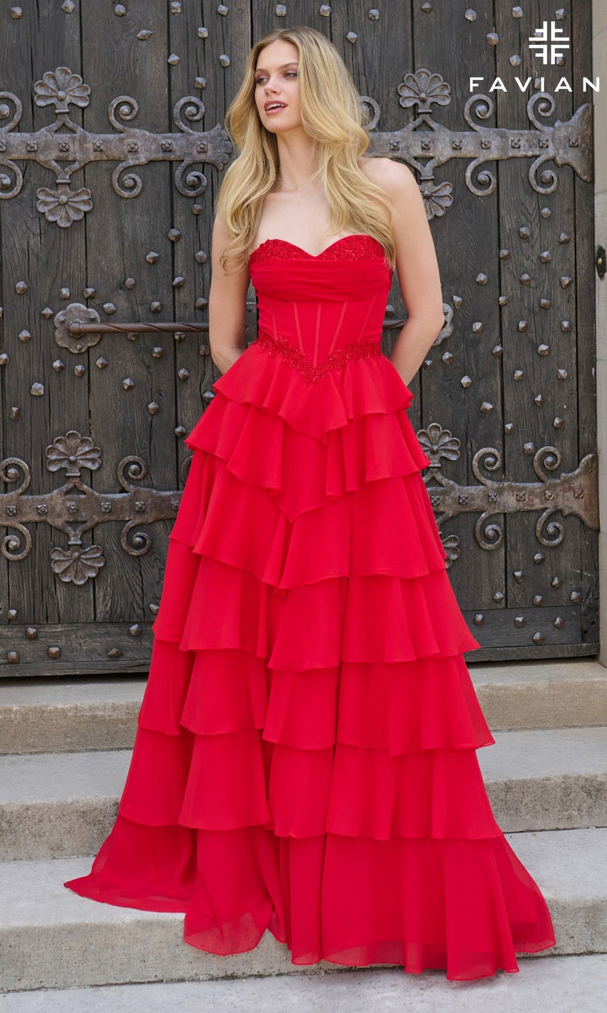 A woman stands before an ornate wooden door, wearing the Faviana 11460 long prom dress with a strapless fitted corset bodice and a flowing tiered ruffle skirt.
